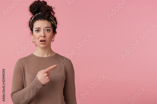 Confused girl with dark curly hair bun. Wearing headband, earrings and brown sweater. Has make up. Watching at the camera and pointing finger to the right at copy space, isolated over pink background
