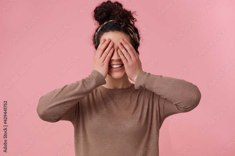 Playful girl, happy looking woman with dark curly hair bun. Wearing headband and brown sweater. Has make up. Emotion concept. Close eyes with palms. Stand isolated over pastel pink background Playful girl, happy looking woman with dark curly hair bun. Wearing headband and brown sweater. Has make up. Emotion concept. Close eyes with palms. Stand isolated over pastel pink background