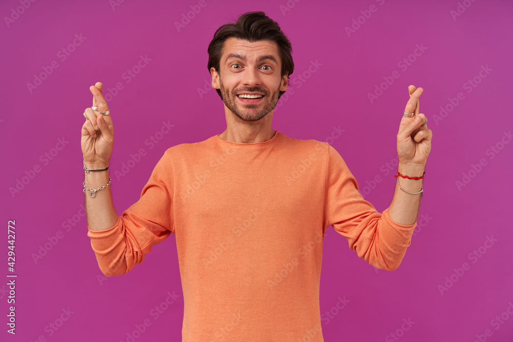Happy looking guy with brunette hair and bristle. Wearing orange sweater with rolled up sleeves. Has bracelets and rings. Keeps fingers crossed. Watching at the camera isolated over purple background