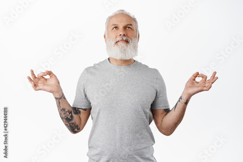 Peaceful senior man feel zen, meditating and resting, breathing calm and looking up, smiling satisfied, calming down, holding hands in yoga mudra signs, standing over white background