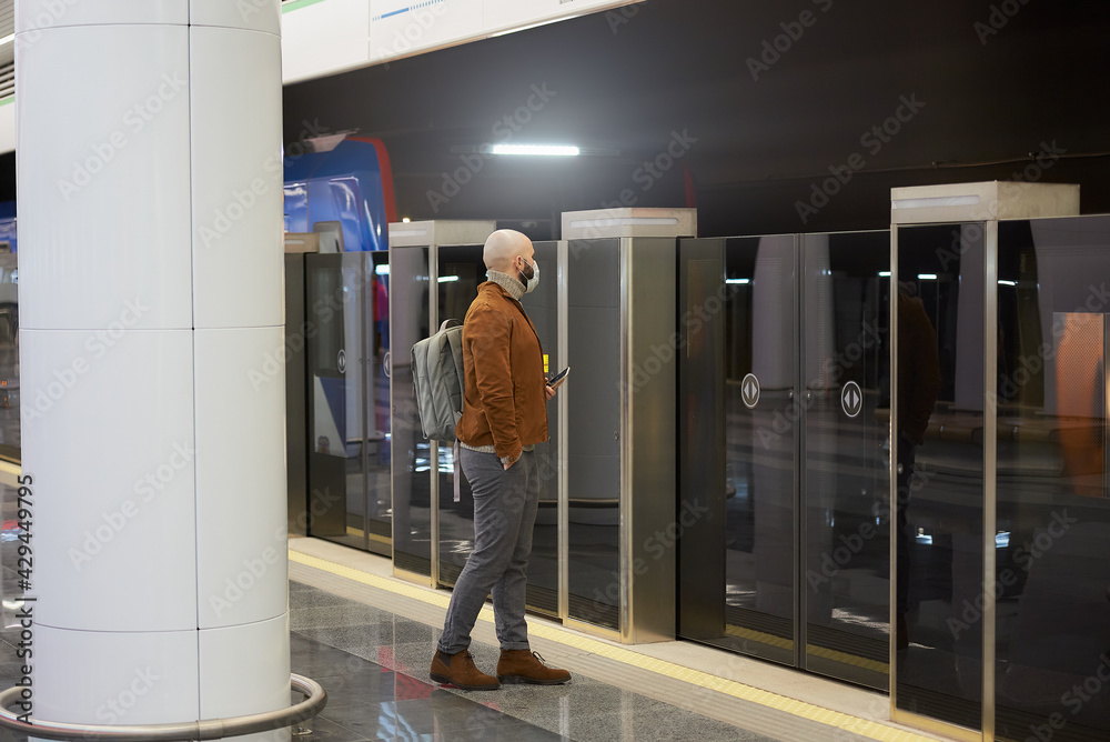 A man in a face mask is holding a smartphone while waiting for a subway train. A man in a face mask is holding a smartphone while waiting for a subway train.