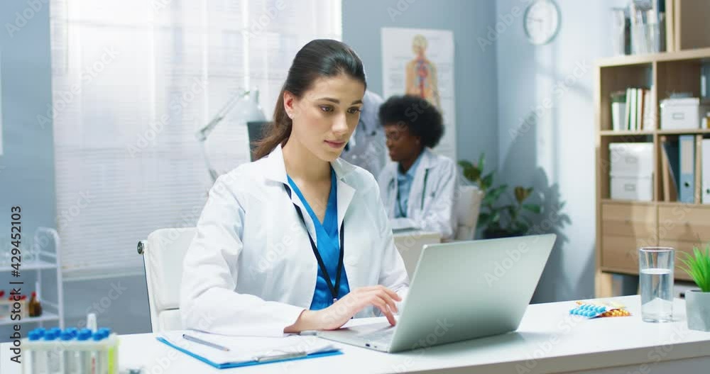 Caucasian young beautiful professional general practitioner woman working in cabinet texting and browsing online on laptop sitting at desk in hospital at workplace. Healthcare, Portrait concept Caucasian young beautiful professional general practitioner woman working in cabinet texting and browsing online on laptop sitting at desk in hospital at workplace. Healthcare, Portrait concept
