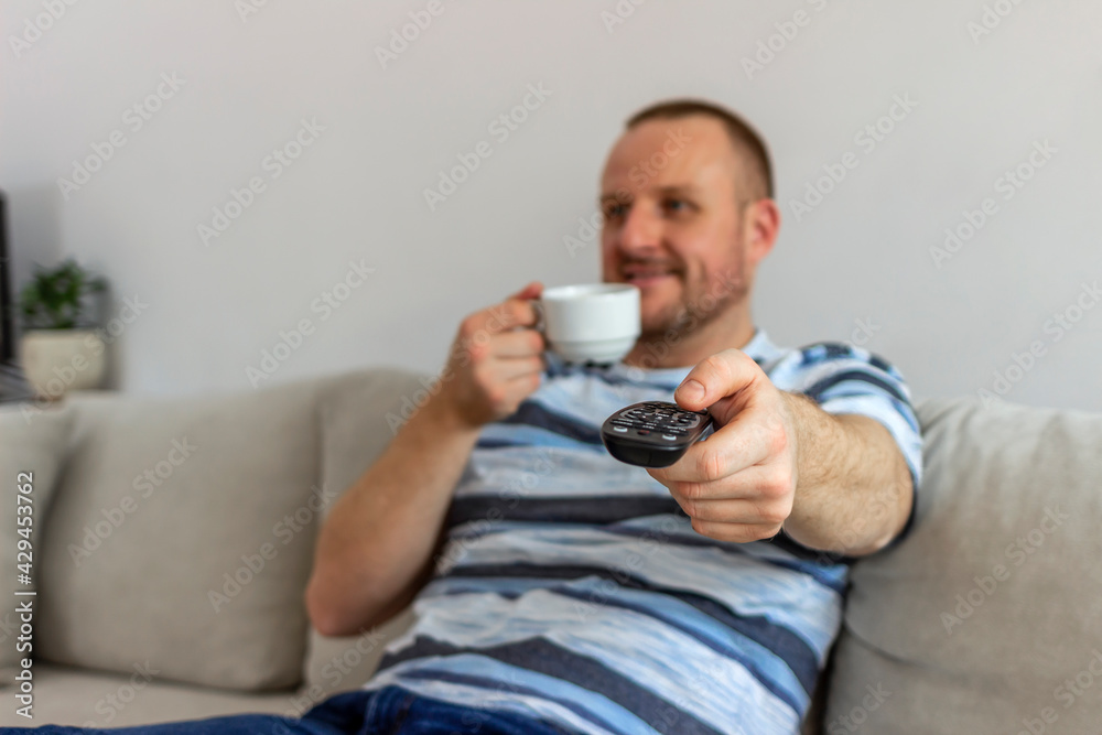 Selective focus on a smiling middle aged man with coffee and remote control at home. Photo of a cheerful bearded young man sitting on couch, using TV remote control looking for another movie.