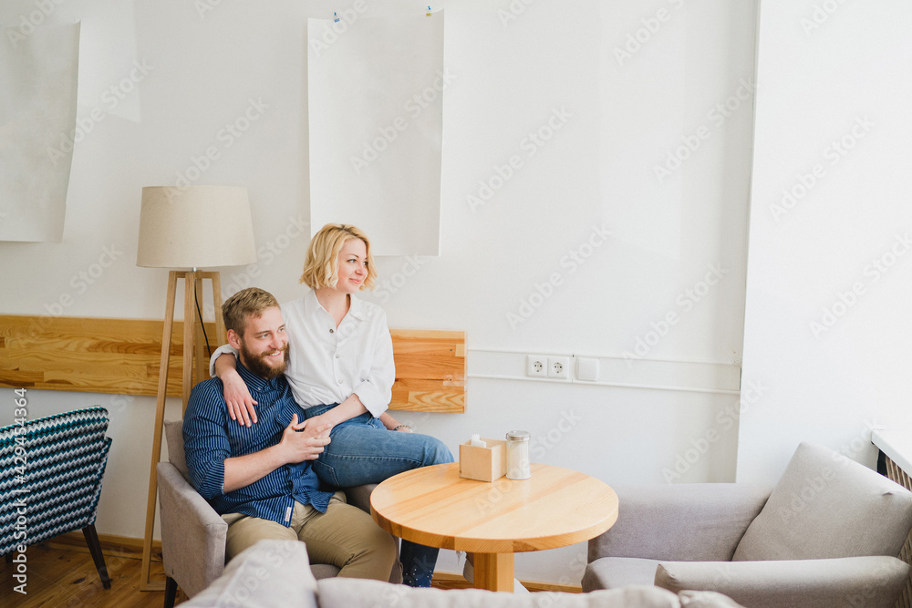 Fototapeta premium Romantic and happy caucasian couple in casual clothes sitting together in an armchair in cozy light space. Love, relationships, romance, happiness concept. Man and woman holding hands and smiling.
