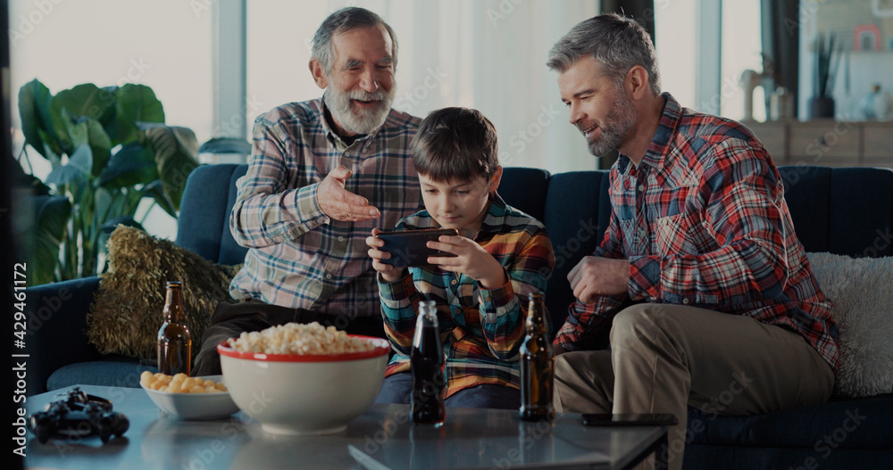 Three family people of father and son with elderly grandpa using mobile phone playing online videogame racing exciting together supporting the child. Three family people of father and son with elderly grandpa using mobile phone playing online videogame racing exciting together supporting the child.