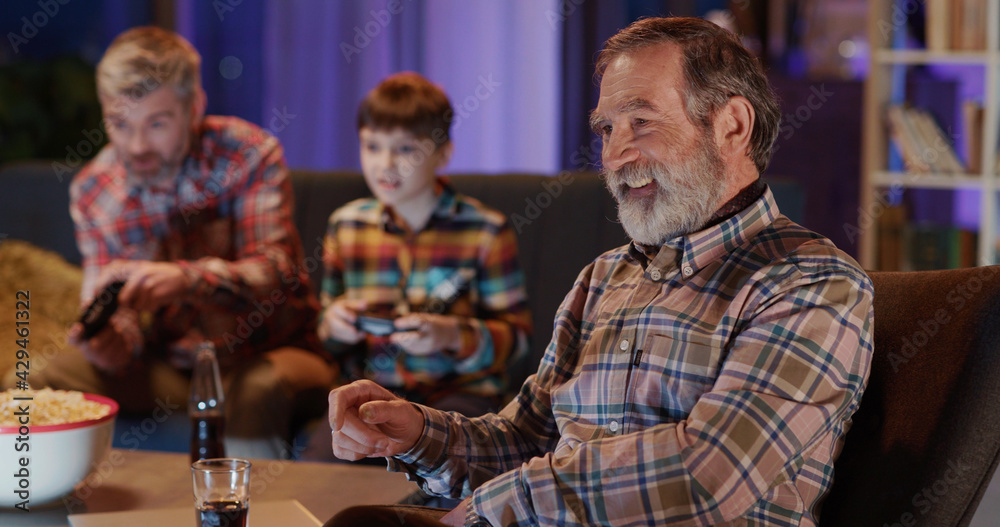 Cheerful caucasian mature adult sitting by television cheering for his son and grandson playing football videogame together.