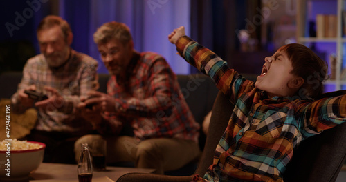 Happy caucasian teenage boy cheering near television watching his father and grandpa playing computer competition game on console.