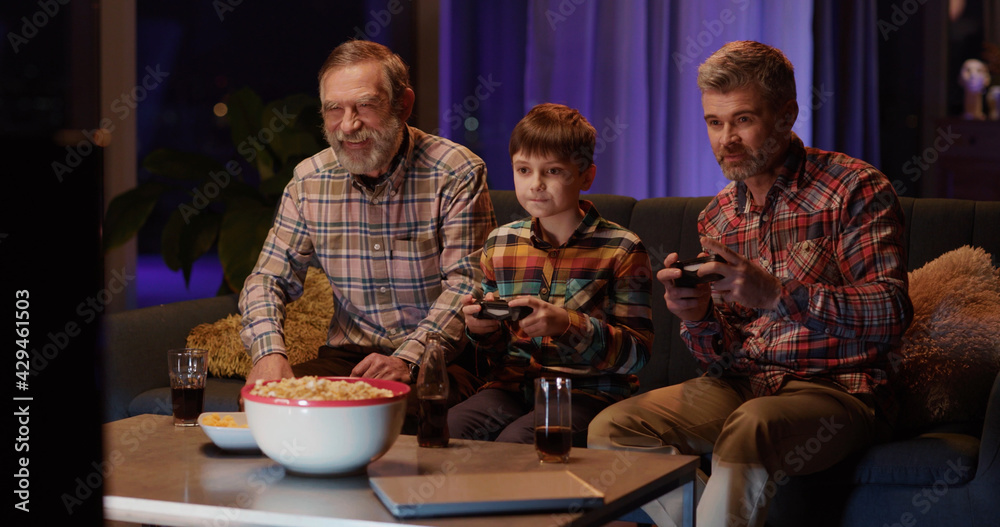 NEW YORK - 19 May 2018: Family of three playing computer games on console together. Multi-generation trio father son and grandpa having fun at home weekend night. NEW YORK - 19 May 2018: Family of three playing computer games on console together. Multi-generation trio father son and grandpa having fun at home weekend night.