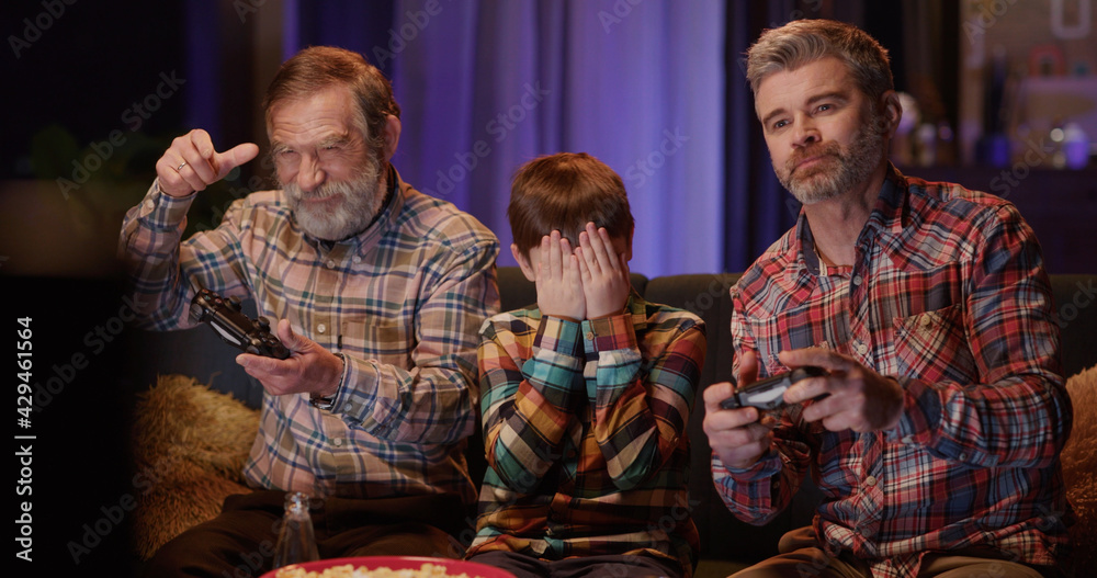 Multi-generational caucasian family of three playing console computer online video games together and eating popcorn snacks. Home entertainment.