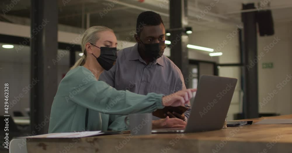 Diverse male and female business colleagues wearing masks in discussion at work using laptop Diverse male and female business colleagues wearing masks in discussion at work using laptop