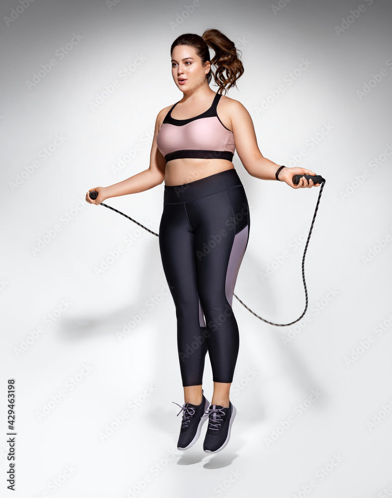 Fototapeta premium Sporty girl with skipping rope in motion. Photo of model with curvy figure in fashionable sportswear on grey background. Dynamic movement. Sports motivation and healthy lifestyle