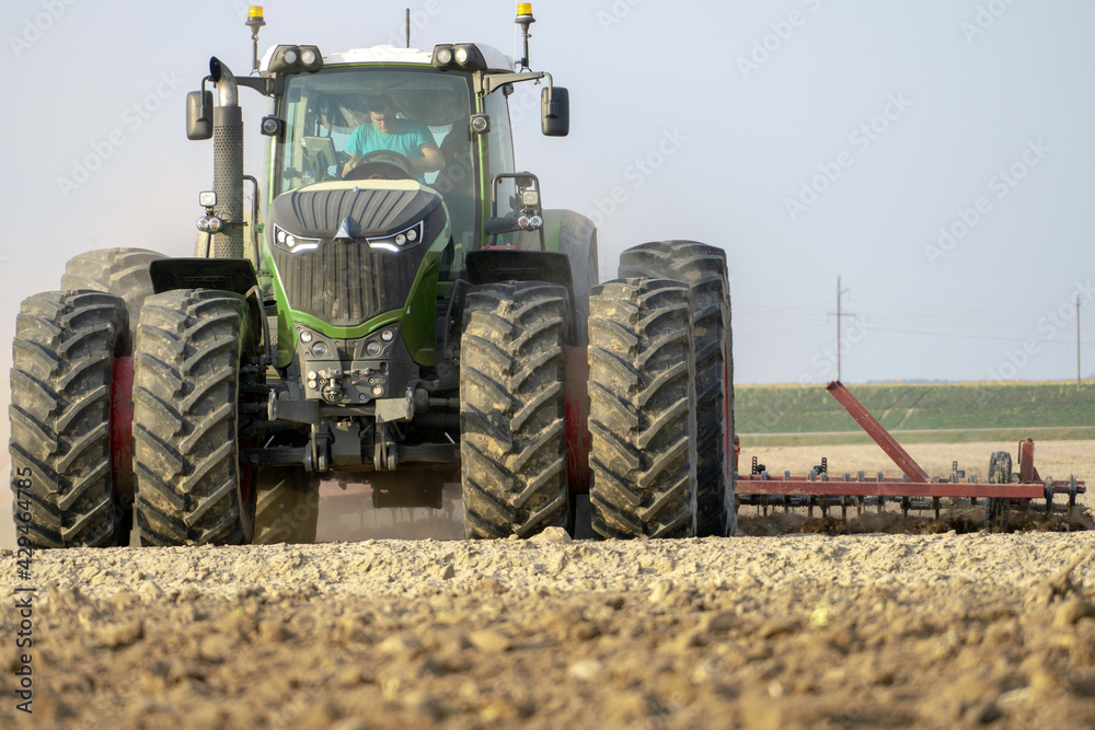 Fototapeta premium A large tractor close-up with a plow working in a field on a summer day against a clear blue sky. Tillage. A professional tractor driver does his job. The beginning of sowing works.