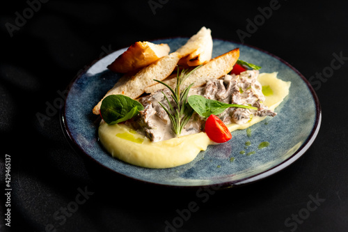 Meat in a creamy sauce with mashed potatoes and bread toast, painted with spinach leaves and cherry tomatoes, on a gray plate on a black background.