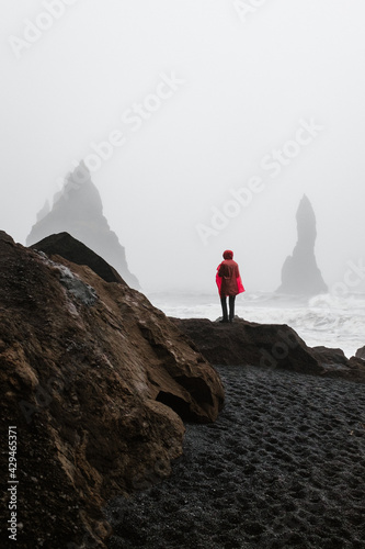 Person in red raincoat standing and looking at the ocean in black sand beach in Iceland