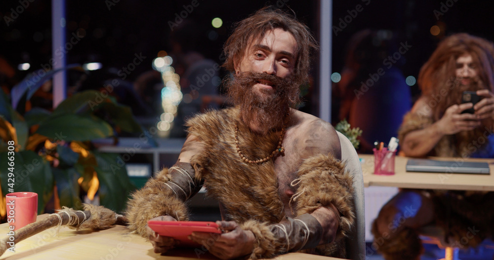 Indoor Portrait of Furry Nomadic Young Caveman with Long Beard Covered Animal Fur Using Tablet Technology Interacting Modern World Working in Office.