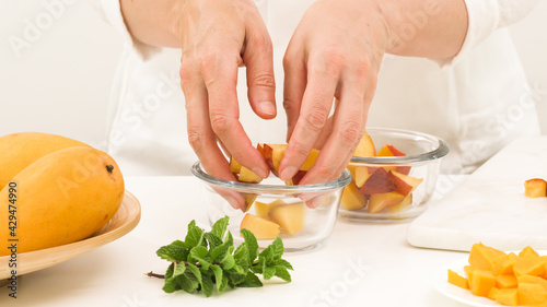 Chopped mangoes and peaches in a glass bowls, woman hands, white background. Fresh fruit salad, delicious dessert