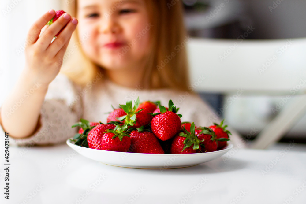 Cute little girl eating fresh strawberry in the kitchen. Healthy vitamin snack for kids. Ripe fresh berries. Harvest season. Natural vitamins . Cute little girl eating fresh strawberry in the kitchen. Healthy vitamin snack for kids. Ripe fresh berries. Harvest season. Natural vitamins .