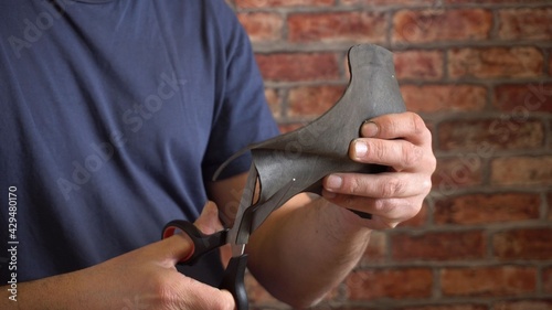 A man carves a fragment out of dark rubber. Scissors in hand. Homemade product. Without a face. Blue T-shirt. Against the background of a brick wall. Close-up.