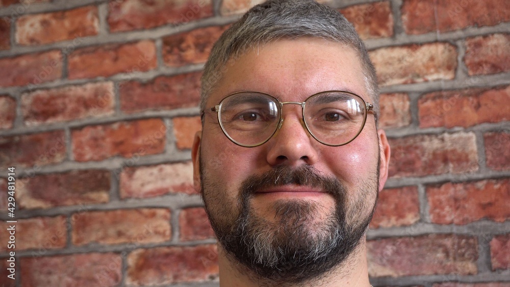 Bearded man in round retro glasses. Looks straight. Short haircut. Blue T-shirt. Poor eyesight. Against the background of a brick wall. Close-up. Bearded man in round retro glasses. Looks straight. Short haircut. Blue T-shirt. Poor eyesight. Against the background of a brick wall. Close-up.