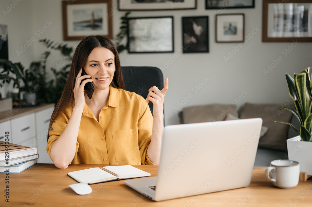 Smiling business woman talking on a mobile phone and using a laptop while working at home, looking at the screen, reading a message, a young female employee advises a client, telephone conversations Smiling business woman talking on a mobile phone and using a laptop while working at home, looking at the screen, reading a message, a young female employee advises a client, telephone conversations