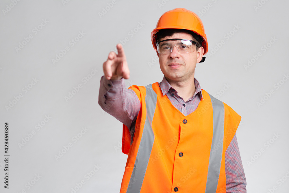 young construction worker in hard hat, protective glasses and overalls on studio background showing with finger direction forward
