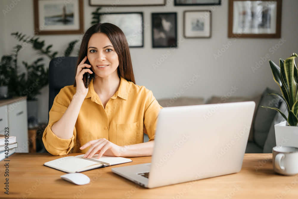 Smiling business woman talking on a mobile phone and using a laptop while working at home, looking at the screen, reading a message, a young female employee advises a client, telephone conversations Smiling business woman talking on a mobile phone and using a laptop while working at home, looking at the screen, reading a message, a young female employee advises a client, telephone conversations