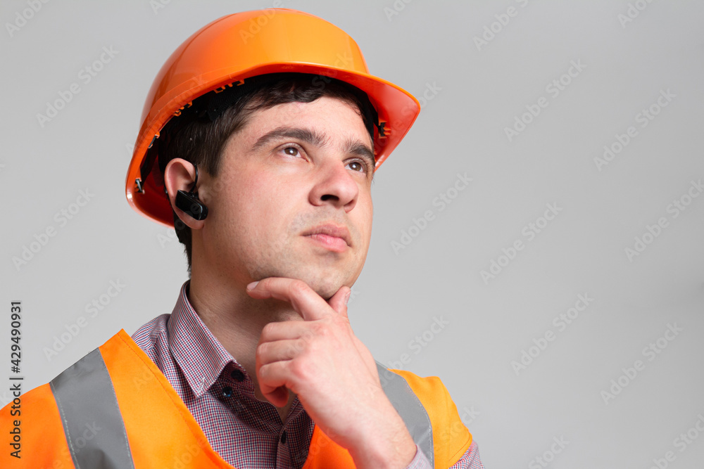 Young engineer architect in hard hat and reflective vest thinking with hand under chin on grey studio background, building industry concept Young engineer architect in hard hat and reflective vest thinking with hand under chin on grey studio background, building industry concept