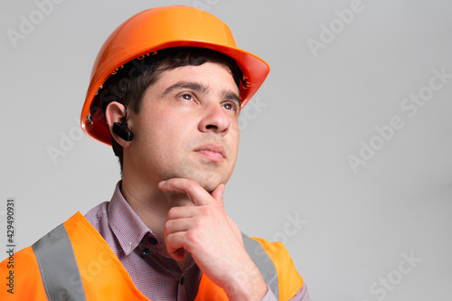 Young engineer architect in hard hat and reflective vest thinking with hand under chin on grey studio background, building industry concept
