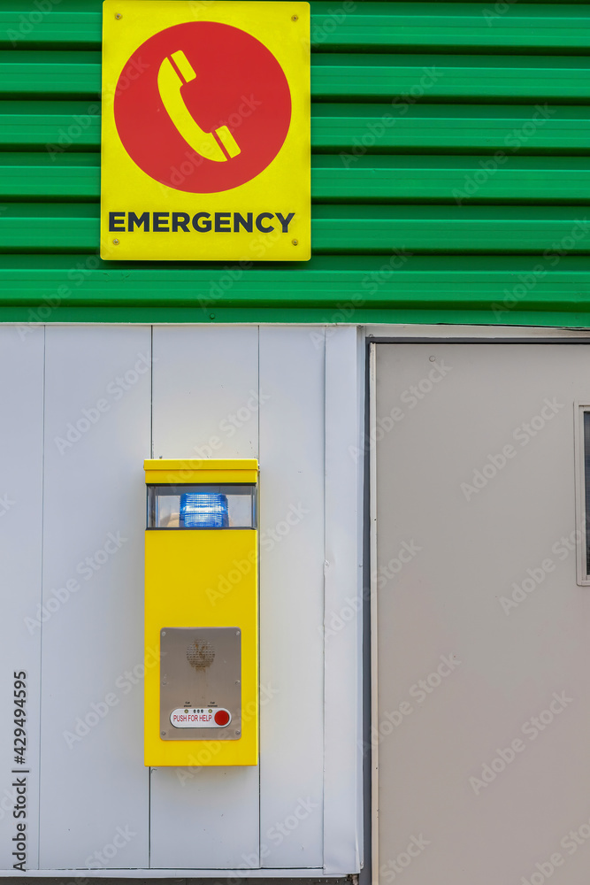 Yellow emergency call box mounted on building exterior, emergency sign, green metal siding, steel door, nobody
