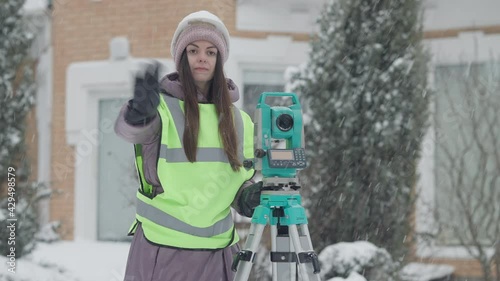 Middle shot of confident young woman using theodolite outdoors on snowy winter day. Skilled Caucasian topographer gesturing adjusting topographic equipment. Industrial concept