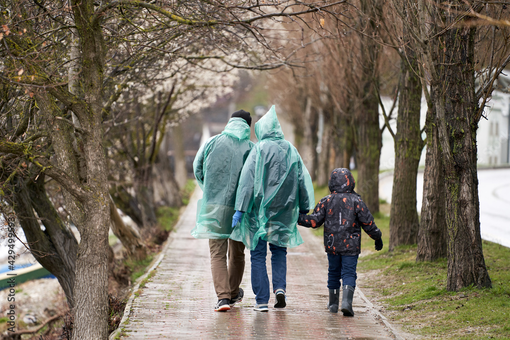Fototapeta premium A family of three strolls down an alley in the rain. The parents are wearing disposable raincoats, the child is wearing a jacket and rubber boots. Selective focus.