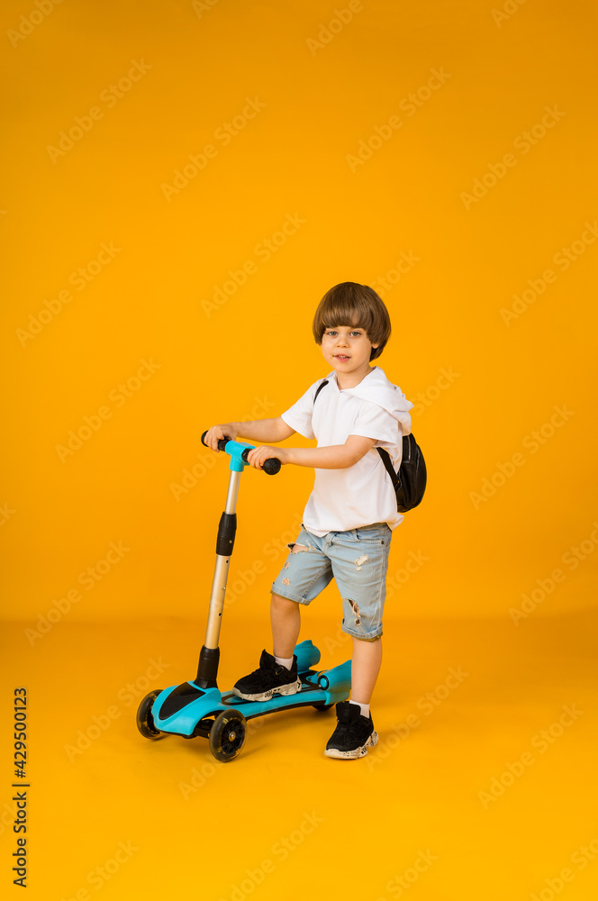 small boy stands with a scooter on a yellow background with space for text. Sports for children small boy stands with a scooter on a yellow background with space for text. Sports for children