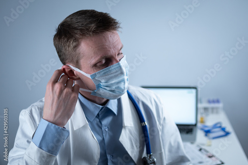 Exhausted young doctor in lab coat takes off mask and glasses, rubs eyes, has headache from fatigue and overexertion from working in heavy shift in hospital. Doctor looks tiredly at camera in office