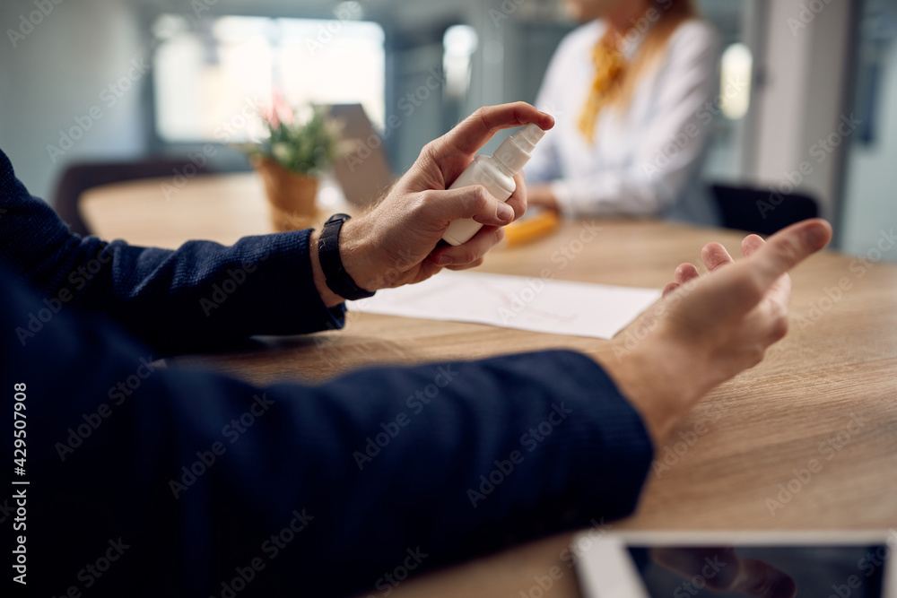 Close-up of entrepreneur spraying hands with disinfectant while working in the office during coronavirus pandemic.