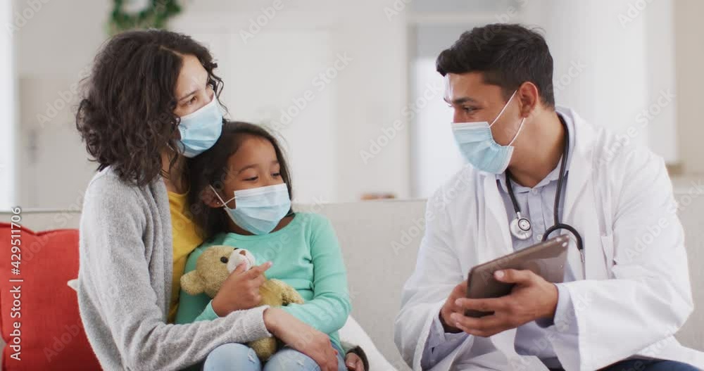 Hispanic male doctor talking to mother and daughter at home, all wearing face masks