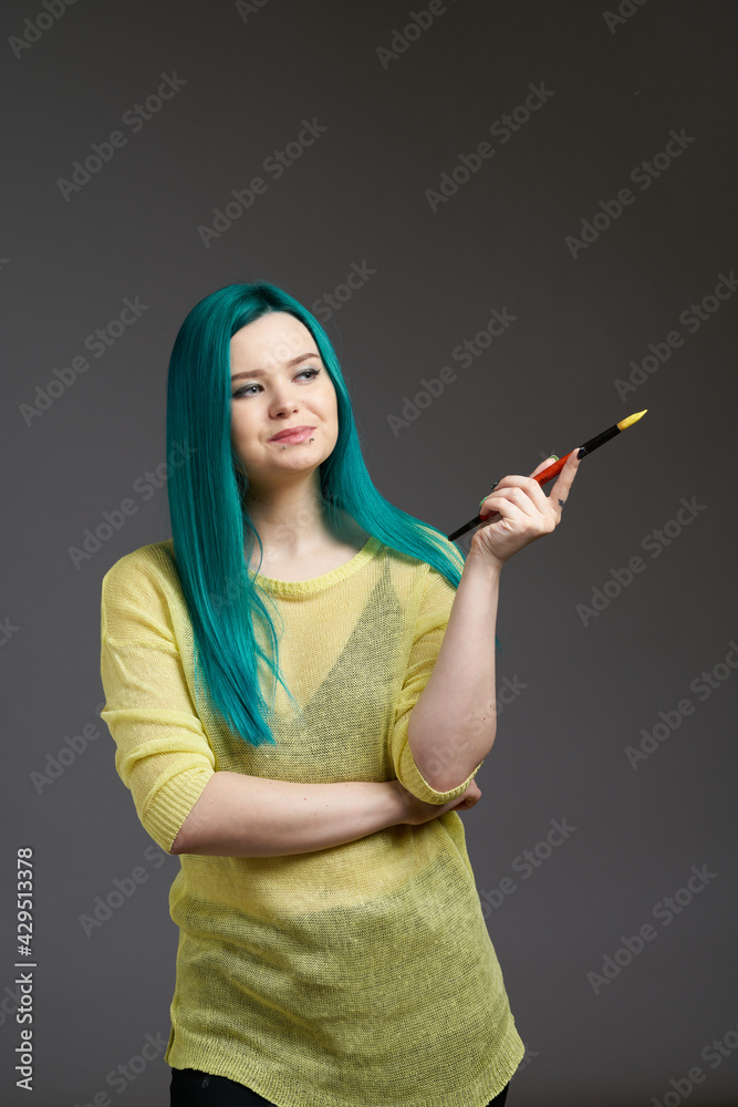 The girl artist holds a brush with paint. Woman with blue green hair in a yellow sweater isolated on a grey background. Bright colored appearance of a creative person The girl artist holds a brush with paint. Woman with blue green hair in a yellow sweater isolated on a grey background. Bright colored appearance of a creative person