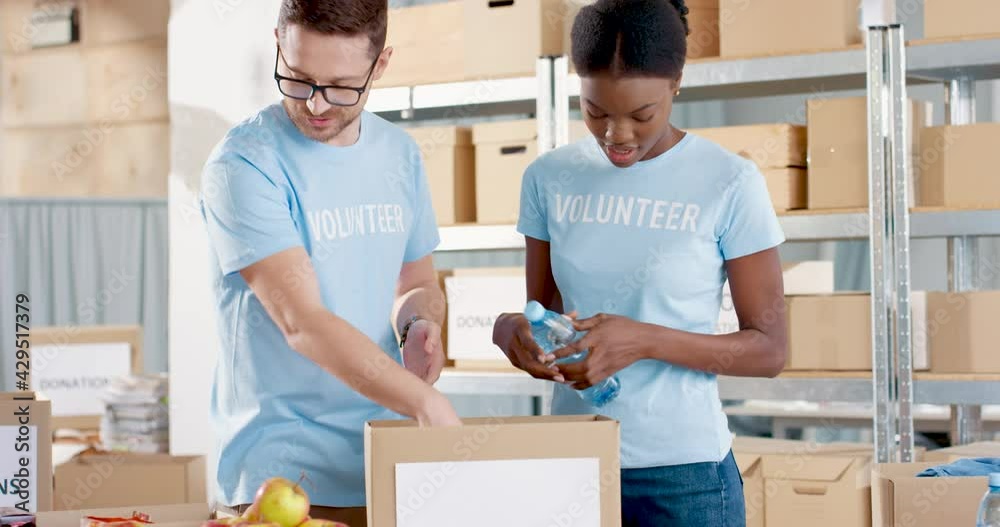 Close up of young happy mixed-race handsome guy and beautiful woman social workers working in charity organization packing food products preparing box with donations. Volunteering and donating Close up of young happy mixed-race handsome guy and beautiful woman social workers working in charity organization packing food products preparing box with donations. Volunteering and donating