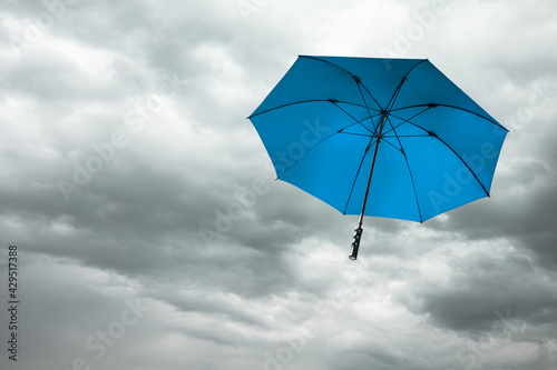 Blue umbrella fly over dark grey cloudy sky with storm wind in rainy season, depth of field. Parasol blow away by heavy windy weather into dramatic cloudscape, overcast clouds background.