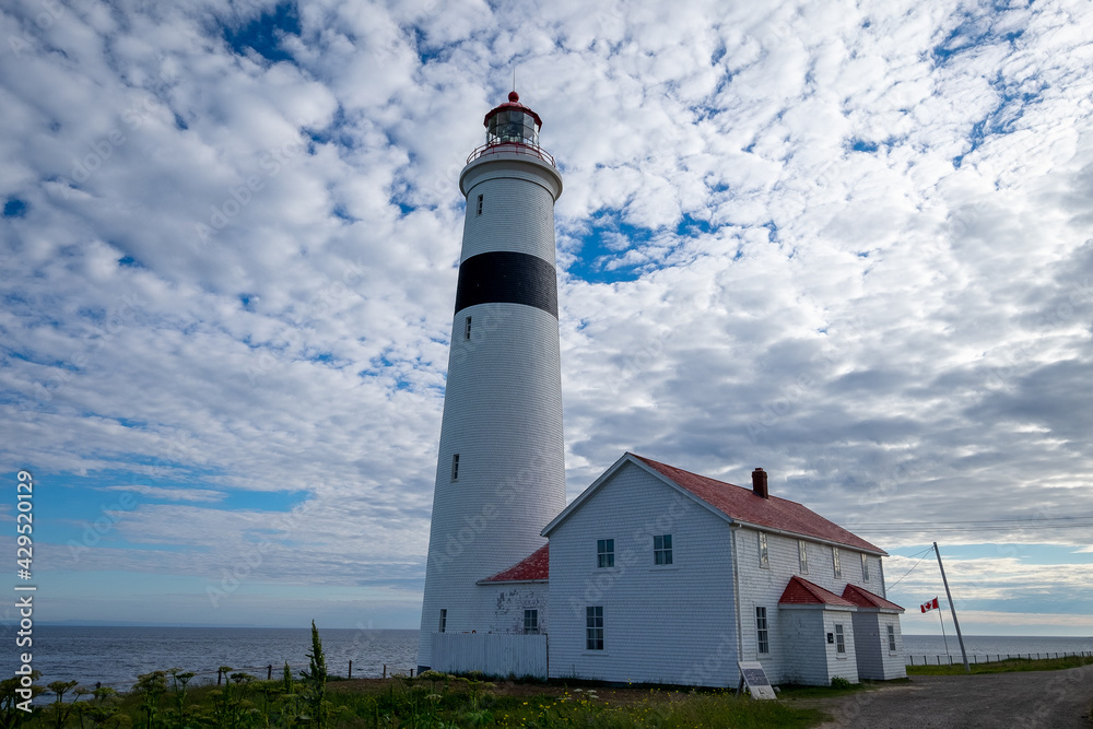 Point Amour Historic Lighthouse on the edge of the Atlantic Ocean. The blue sky is hidden by fluffy clouds.A building with two entrances and a red roof is joined to tower which has a large lamplight. Point Amour Historic Lighthouse on the edge of the Atlantic Ocean. The blue sky is hidden by fluffy clouds.A building with two entrances and a red roof is joined to tower which has a large lamplight.