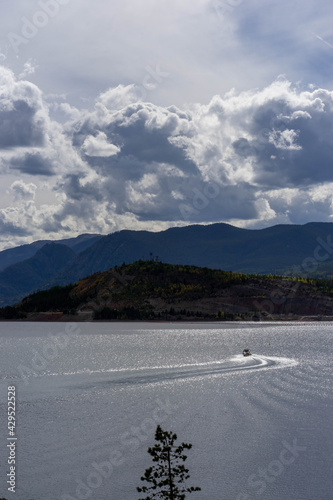 Hillside and mountains with some fall foliage behind sailboat on Lake Dillon, Colorado, on partly cloudy late summer day with some sun shining through the clouds
