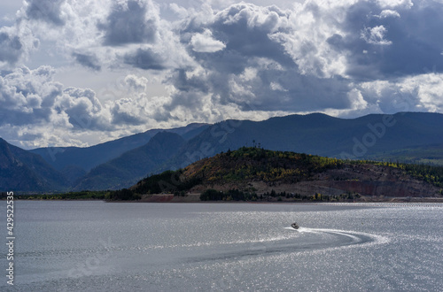Hillside and mountains with some fall foliage behind sailboat on Lake Dillon, Colorado, on partly cloudy late summer day with some sun shining through the clouds