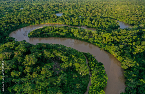 Aerial view of a tropical river meandering through a tropical forest with a canoe going over the river heading to an indigenous community visible by grey tin roofs between the green of the rainforest