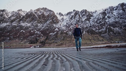 Young man hiking over a beach with sand ripples, beautiful snow-covered mountains in the background, Hoddevik, Norway. photo