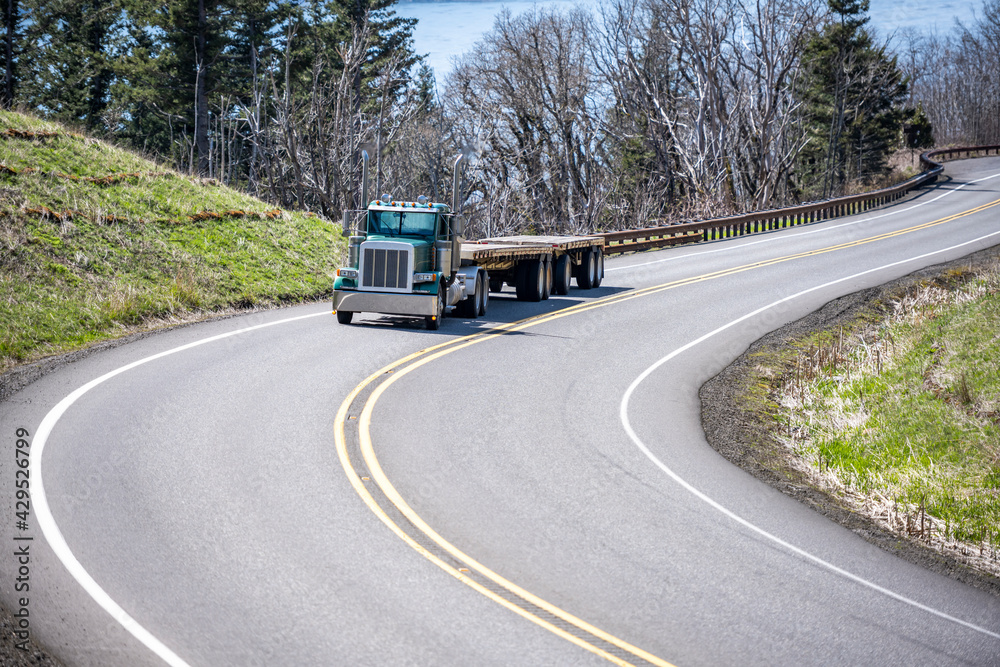 Day cab dark green classic big rig semi truck with two empty flat bed semi trailers running on the winding mountain road at Columbia Gorge Day cab dark green classic big rig semi truck with two empty flat bed semi trailers running on the winding mountain road at Columbia Gorge