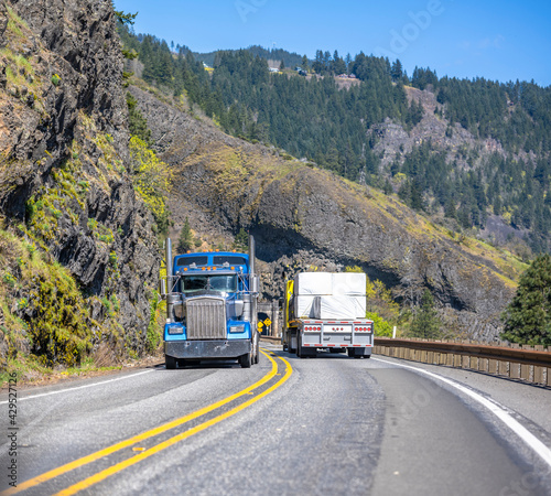 Two different classic loaded big rigs semi trucks with semi trailers moving in opposite directions on the narrow winding road with tunnel and rock on the side at national Columbia Gorge Area