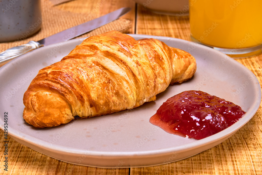 french croissant served with strawberry jam, a glass of orange juice and a cup of coffee, with loaves of bread in the background on a wooden table with a backdrop of white planks. close up