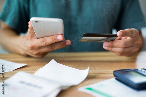Young man wearing wear casual clothes hand holding smart phone and credit card paying bills related to home expenses such as water and electricity bills with calculator bill on wooden office desk.