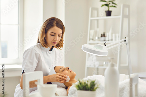 Young cosmetologist or dermatologist cleaning lying womans face with cotton pads in spa salon photo
