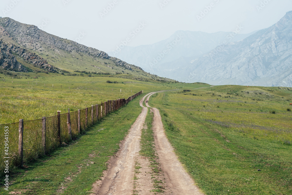 Beautiful green mountain landscape with long dirt road along fence and big mountains in fog. Atmospheric foggy mountain scenery with dirt road among rocks and big mountains. Length road in countryside