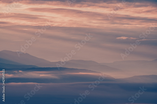 Scenic dawn mountain landscape with lilac low clouds in valley among mountains silhouettes under cloudy sky. Vivid sunset or sunrise scenery with low clouds in mountain valley in pink violet colors.